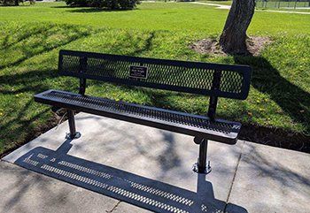 A metal bench with a bronze plaque is seen in a park on a concrete slab