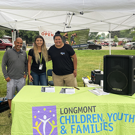 Three smiling people stand under a pop-up tent behind a table that reads "Children, Youth & Families'