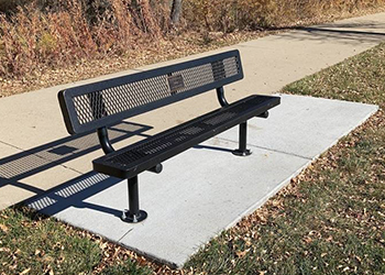 A metal memorial bench is shown on a concert pad in a park