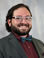 Aren Rodriguez, a middle-aged white man wearing a maroon dress shirt, gray blazer and blue bow tie, sits for a portrait as At Large City Council representative