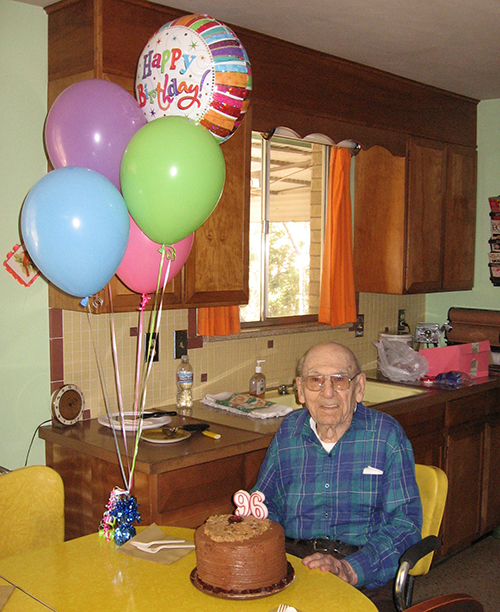 An elderly man seated at a table with a birthday cake and balloons