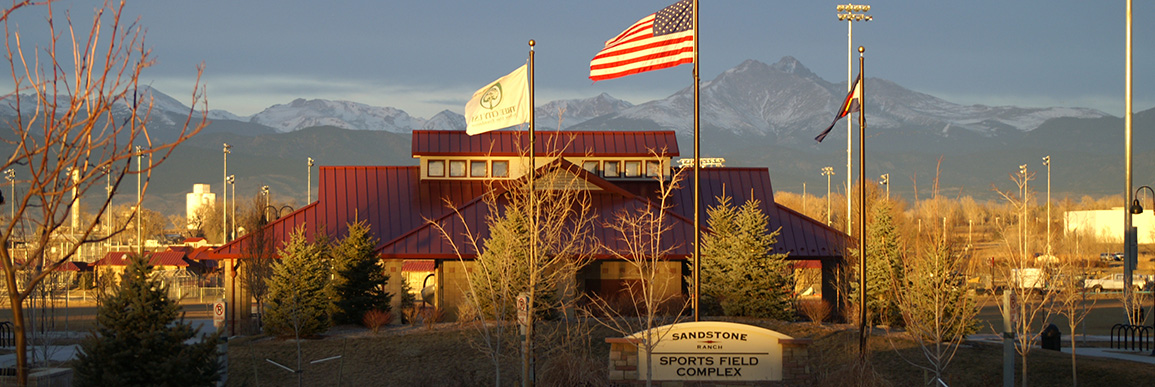 The Sandstone Ranch Sports Complex building and sign, with flags in the wind, in the morning sunlight. A mountain range is in the background.