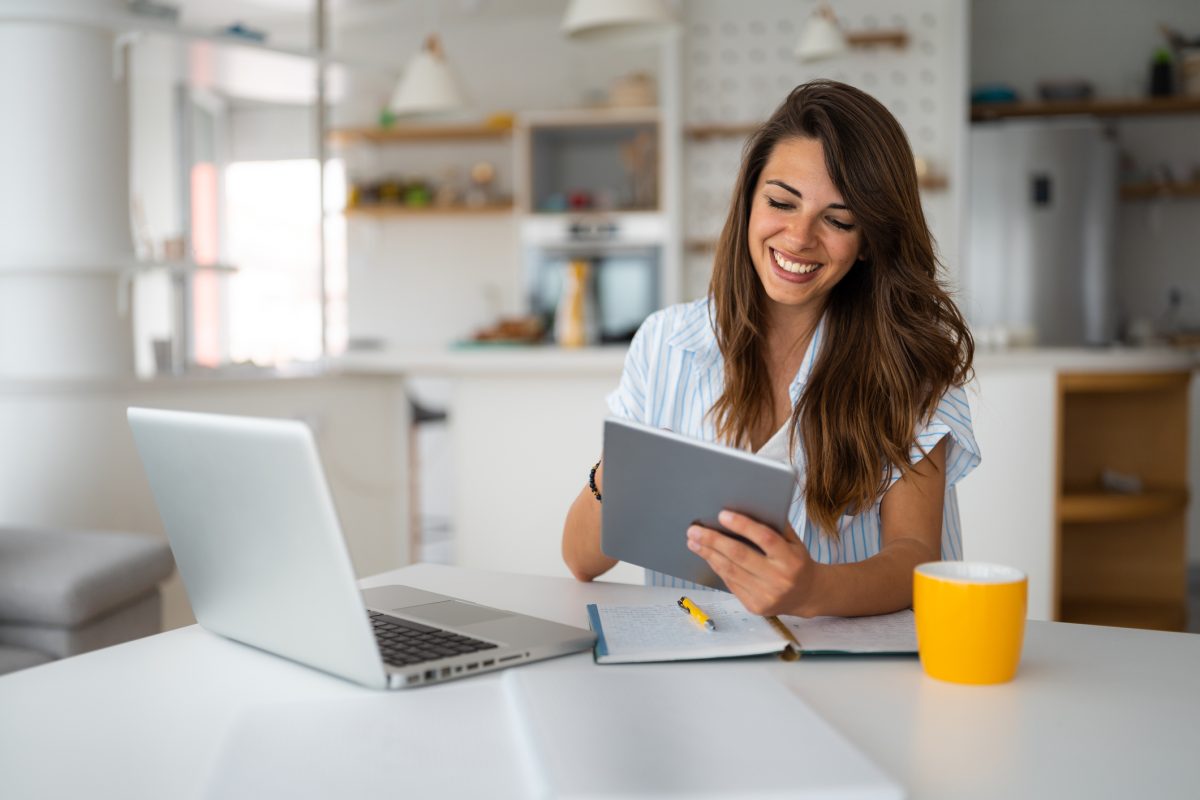 A smiling woman at home reads a digital tablet near her open laptop.